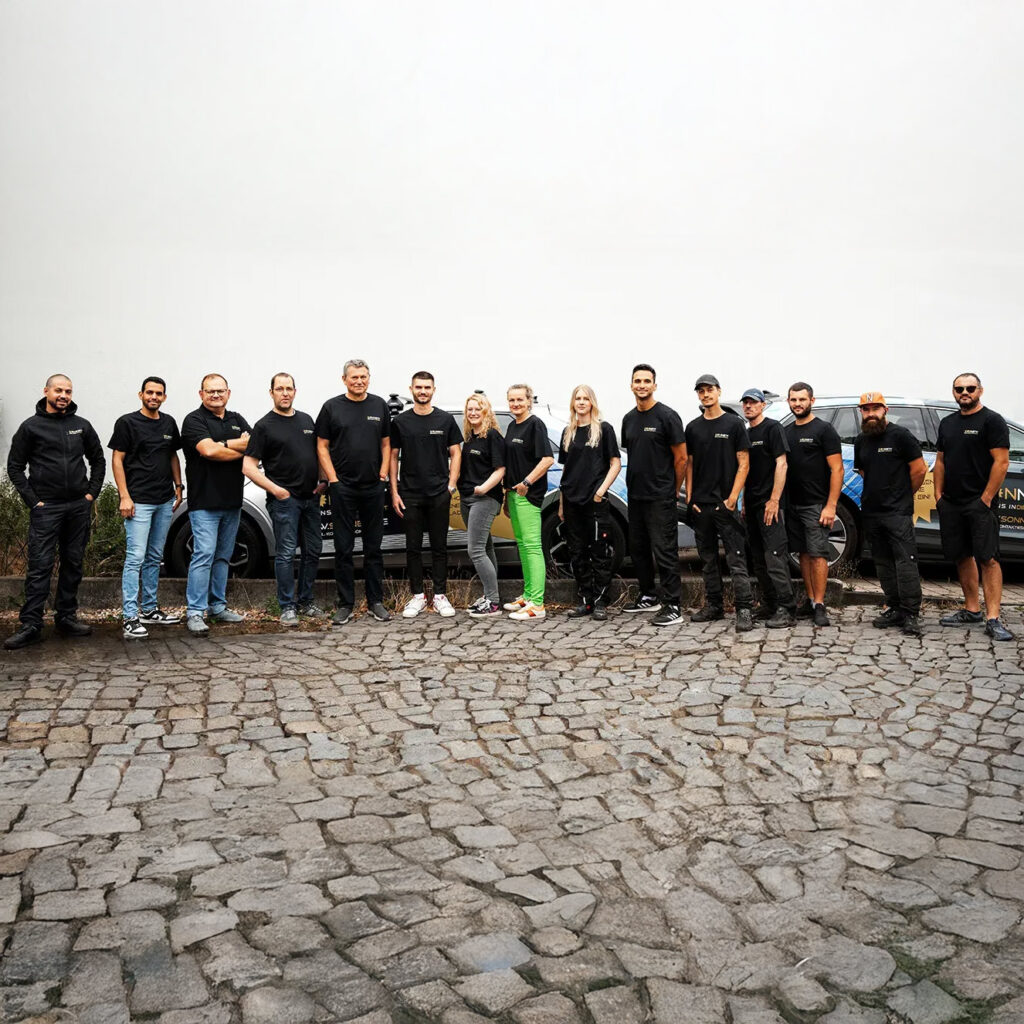 Eine Gruppe von 15 Menschen steht lächelnd auf Kopfsteinpflaster, alle tragen schwarze Kleidung und zwei schwarze Vans sind im Hintergrund sichtbar