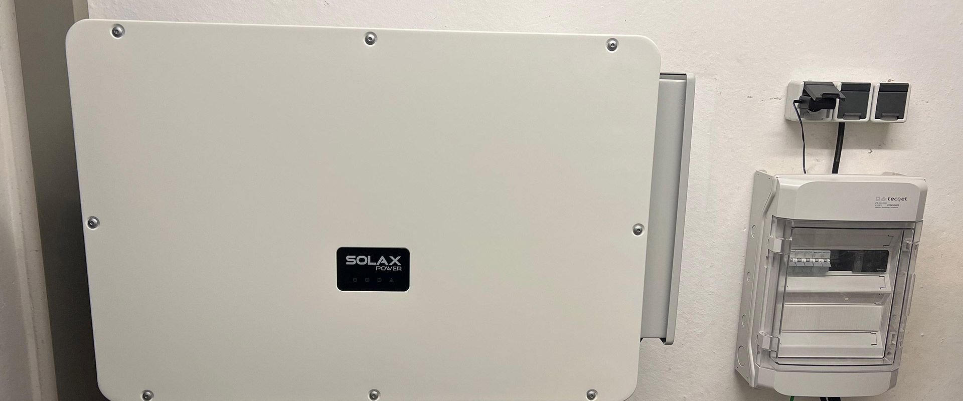 Ein weißes Solax Power Gerät hängt an einer Wand neben einem Sicherungskasten und Steckdosen