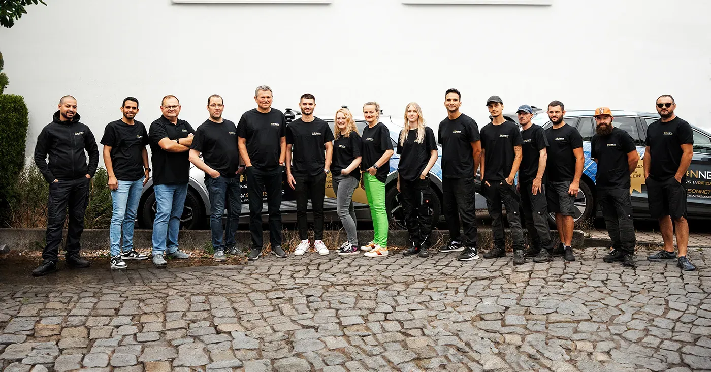 Dreizehn lächelnde Erwachsene in schwarzen Shirts stehen auf Kopfsteinpflaster vor einem Fahrzeug