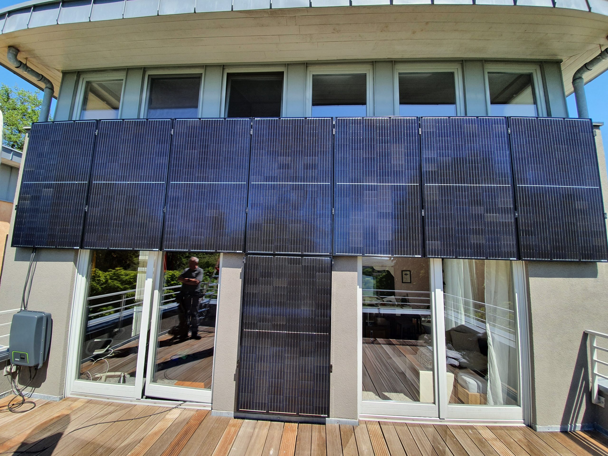 Eine Glasfassade mit mehreren schwarzen Solarpanels und warmer Holzterrasse