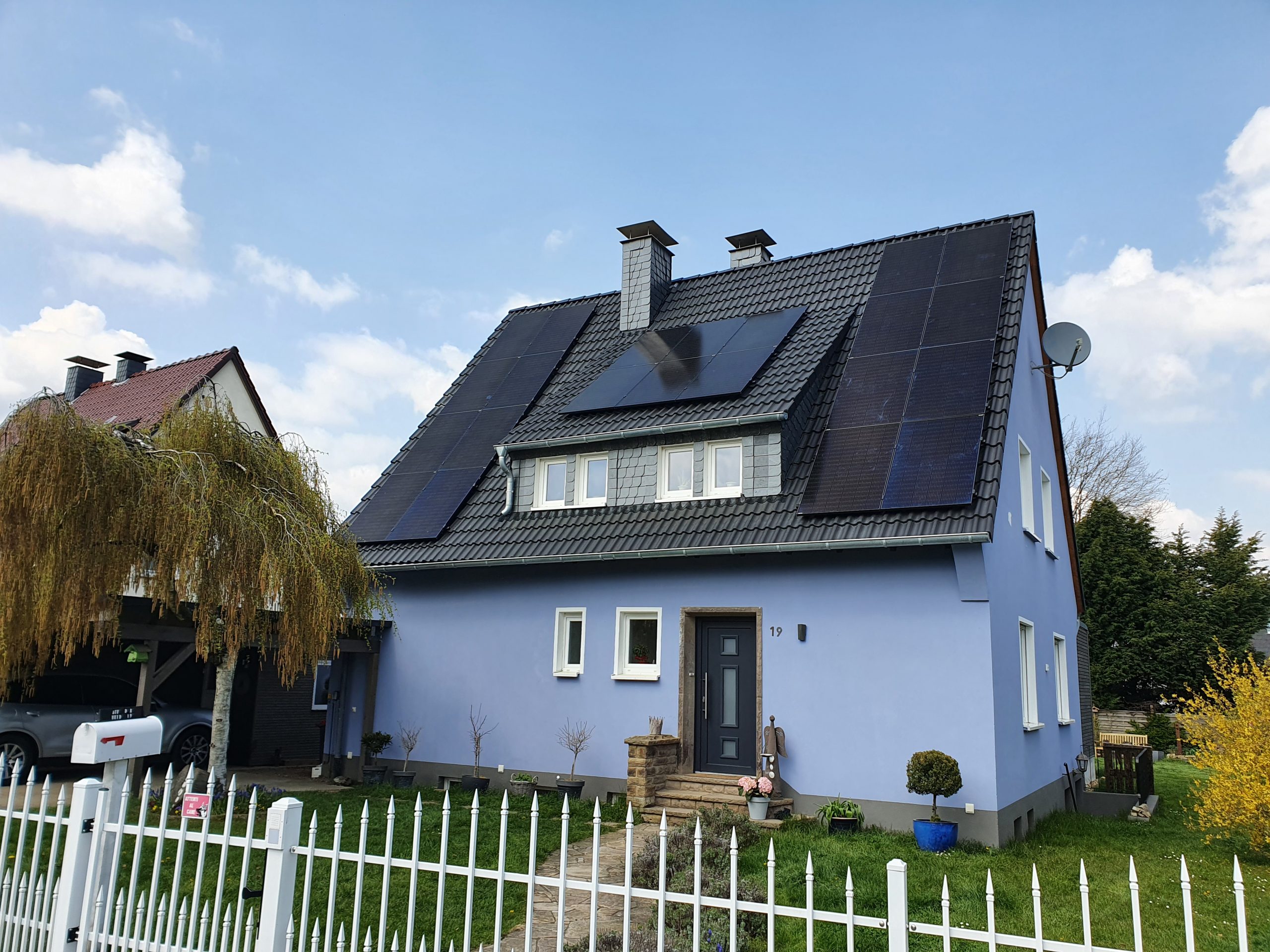 Ein hellblaues Einfamilienhaus mit schwarzen Solarpanels auf dem Dach und gepflegtem Garten