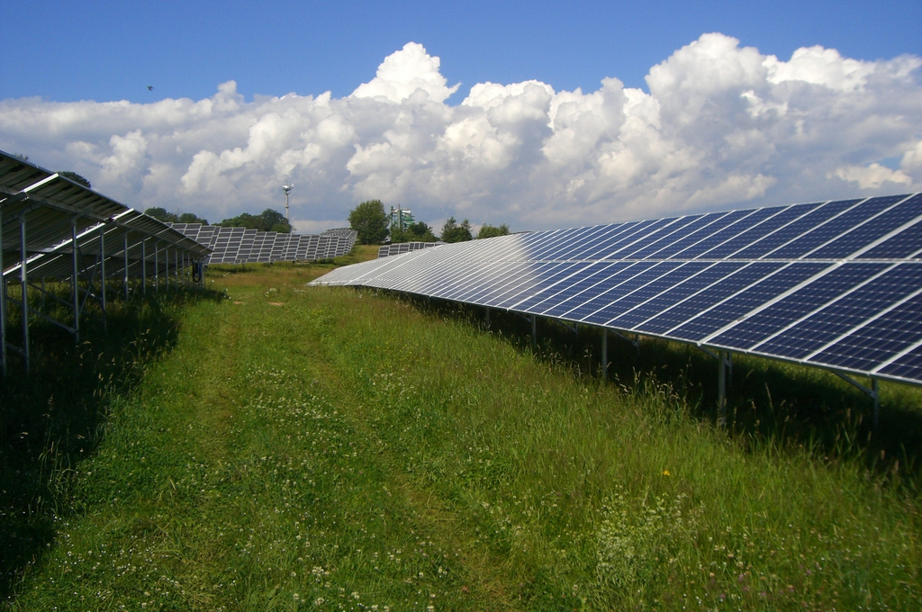 Eine große Solaranlage mit glänzenden blauen Solarpanels steht auf grüner Wiese unter wolkigem Himmel