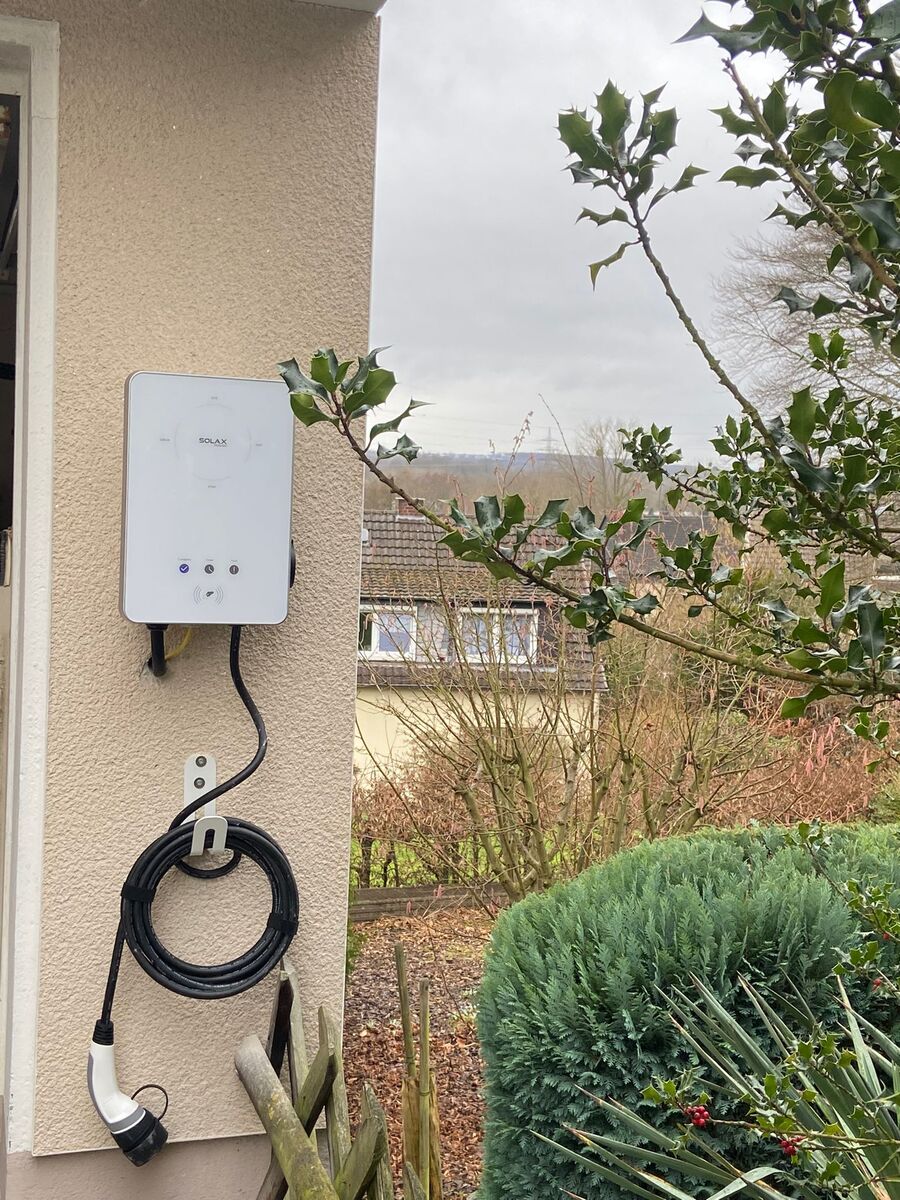 Weiße Wallbox an Hauswand mit Kabel, grauer Himmel und Strauchwerk