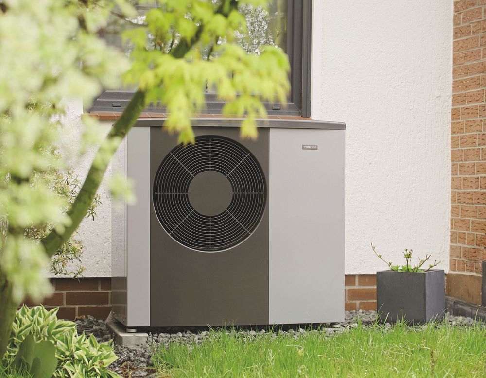 Graue Waermepumpe mit grossem Ventilator neben Hauswand im Gruenen