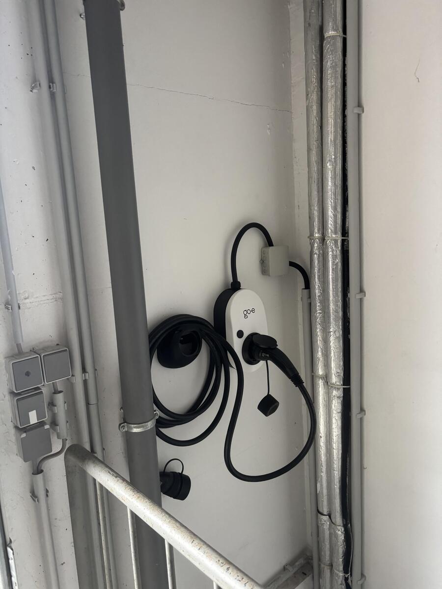 Weisse Ladebox mit schwarzem Kabel an grauer Wand neben Metallrohren