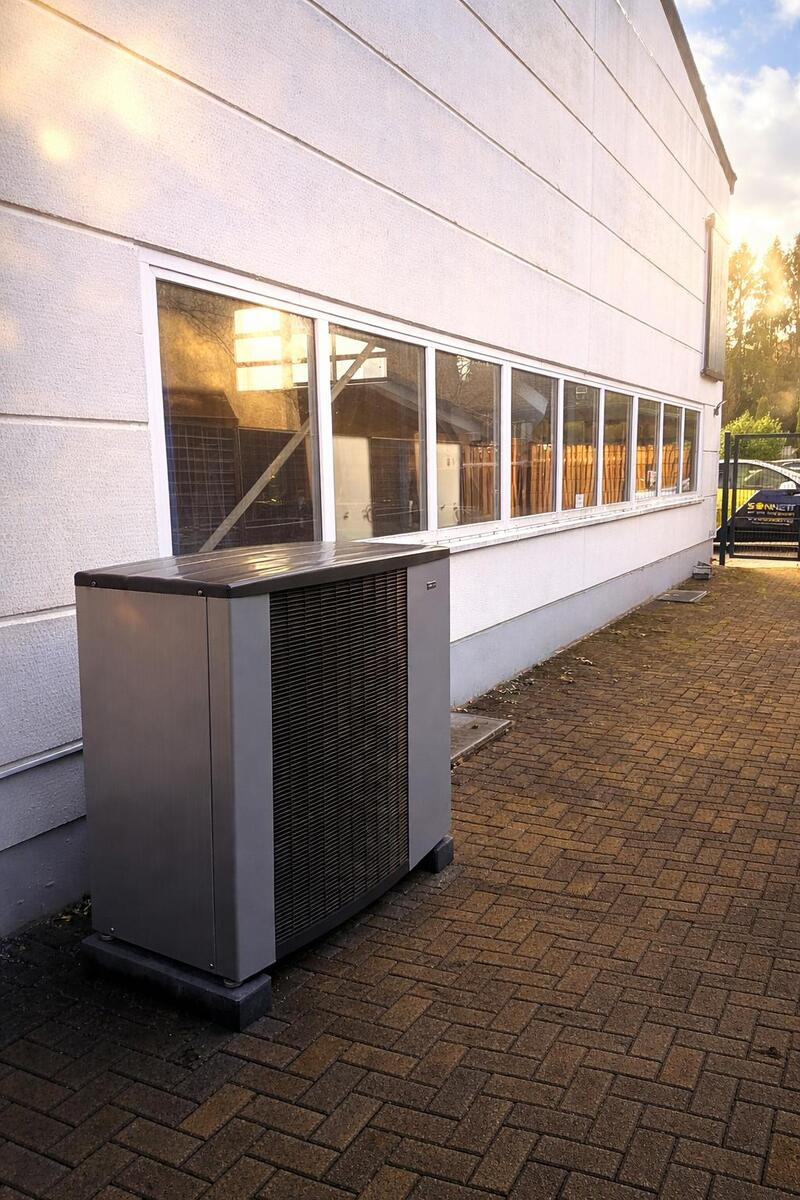 Graues Außengerät an weißer Wand mit Fenstern im Abendlicht, leerer Weg
