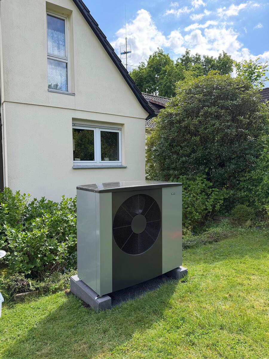 Grüne Wärmepumpe mit schwarzem Lüfter neben Haus im Garten, sonnig