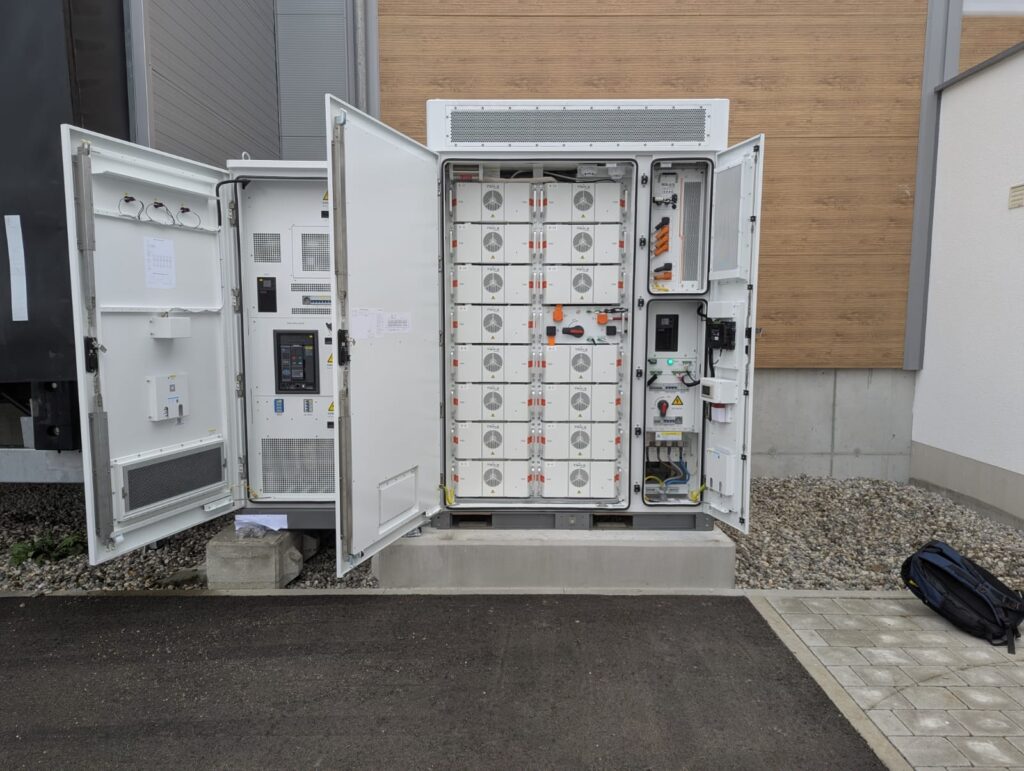 Geoeffneter weisser Batteriespeicherschrank mit Modulen vor einer Holzfassade