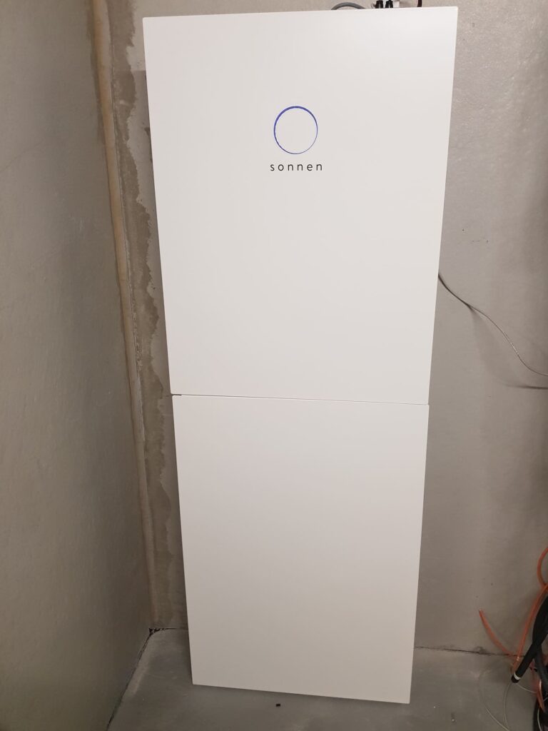 Weisser Sonnen Batteriespeicher mit blauem Logo an einer grauen Kellerwand